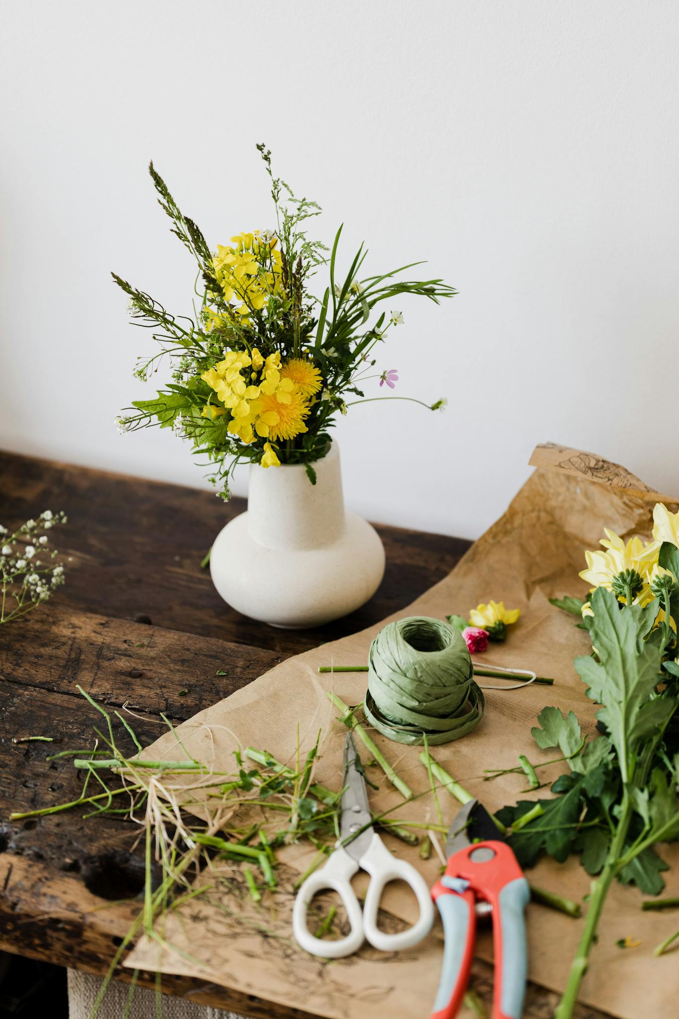 From above of tender yellow flowers in white ceramic vase placed on messy wooden table covered in cut leaves with scissors and pruner
